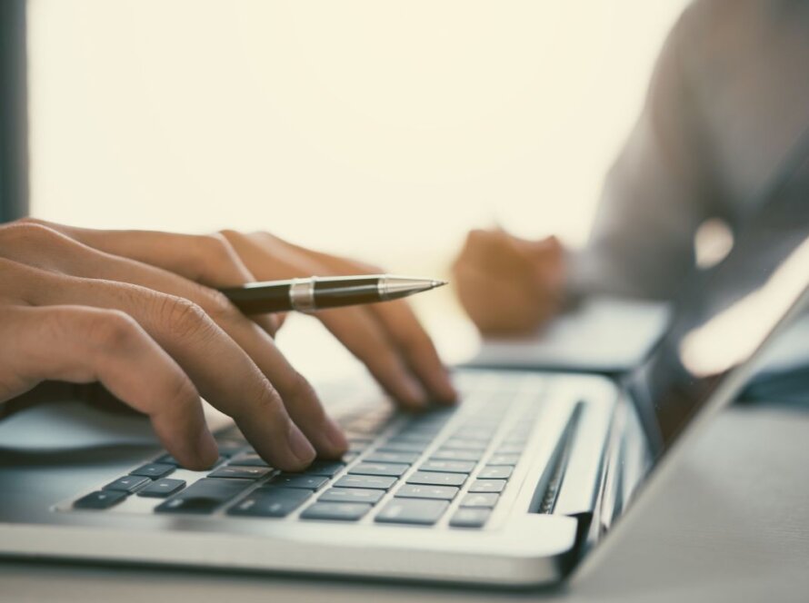 a hand holding a pen while typing on a laptop computer